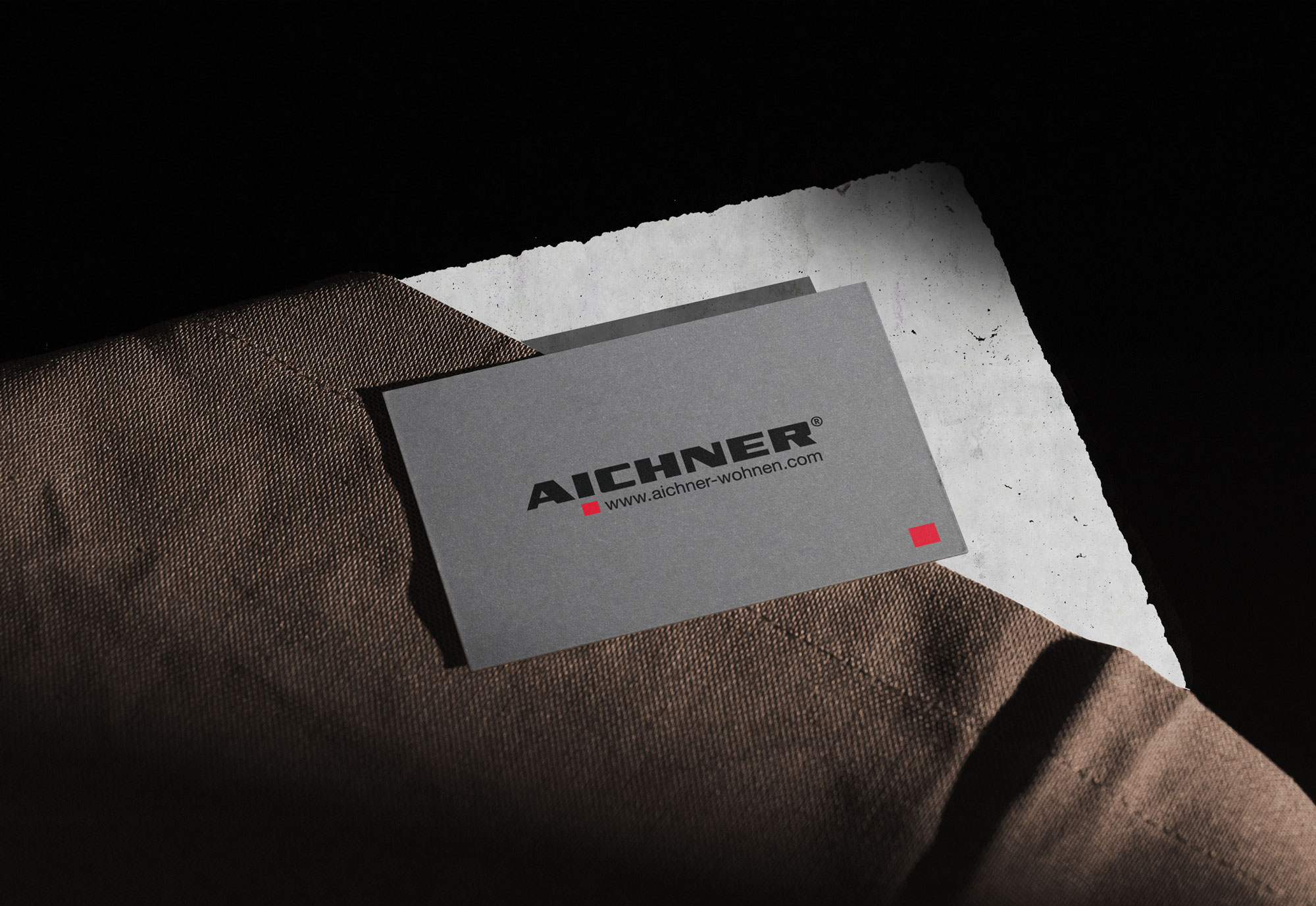 Dunkelgraue Visitenkarte mit dunkelgrauen Strukturpapier und dem Einrichtungshaus Aichner Logo darauf gedruckt darunter ein dunkelbraunes Stück Stoff auf einem Betonpodest mit schwarzen Hintergrund